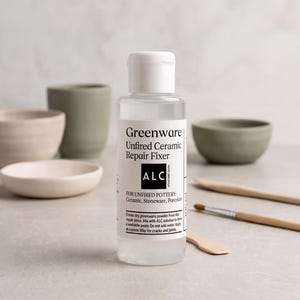 May include: A clear bottle of "Greenware Unfired Ceramic Repair Fixer" with a white cap, next to ceramic bowls and tools. The bottle's label includes instructions for use with ceramic, stoneware, and porcelain. The background features various pottery items.