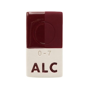 Può includere: Un oggetto rettangolare bicolore, bordeaux nella metà superiore e crema in quella inferiore. La sezione superiore ha una finitura lucida e un'indentazione circolare. La metà inferiore mostra i numeri "0-7" e le lettere "ALC" in bordeaux.