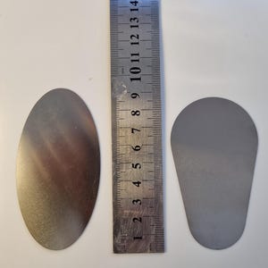 May include: Two metal objects, one oval and one teardrop-shaped, sit next to a ruler. The oval object is approximately 5 inches long. The teardrop object is about 3 inches long. The ruler shows measurements in inches and millimeters.