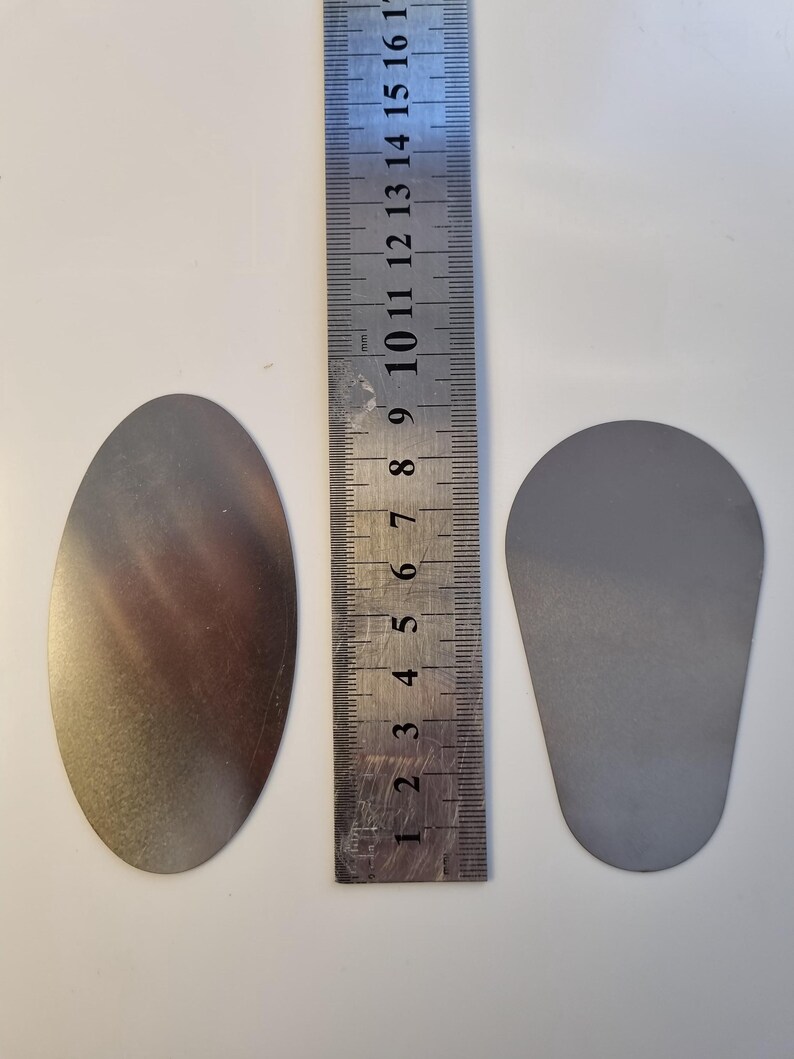 May include: Two metal objects, one oval and one teardrop-shaped, sit next to a ruler. The oval object is approximately 5 inches long. The teardrop object is about 3 inches long. The ruler shows measurements in inches and millimeters.