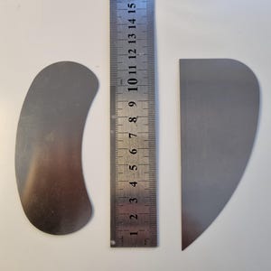 May include: Two metal tools, one curved and one triangular, sit next to a ruler. The ruler shows measurements in centimeters. The curved tool is approximately 4 inches long. The triangular tool is approximately 3 inches long. Both tools are silver.