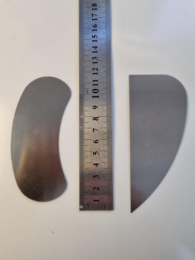 May include: Two metal tools, one curved and one triangular, sit next to a ruler. The ruler shows measurements in centimeters. The curved tool is approximately 4 inches long. The triangular tool is approximately 3 inches long. Both tools are silver.