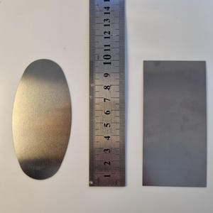 May include: A metal oval and a rectangular metal piece are next to a ruler. The ruler is marked in inches and centimeters. The oval is approximately 3 inches long. The rectangular piece is approximately 2 inches long.