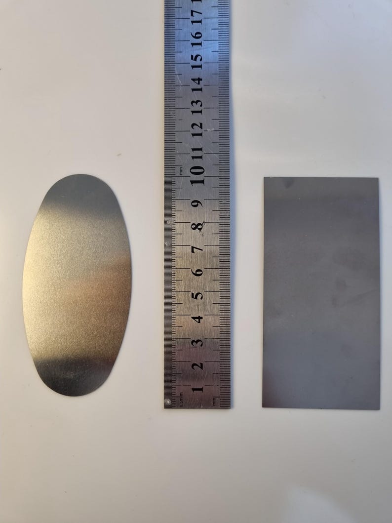 May include: A metal oval and a rectangular metal piece are next to a ruler. The ruler is marked in inches and centimeters. The oval is approximately 3 inches long. The rectangular piece is approximately 2 inches long.