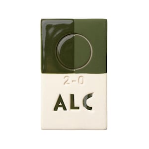 May include: A rectangular ceramic object with a dark green top half and a white bottom half. The top has a circular indentation with a metal ring. The bottom half has the numbers "2-0" above the letters "ALC".