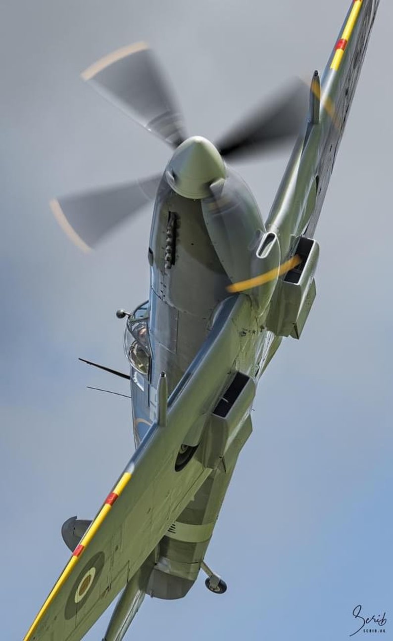 May include: A green and silver Supermarine Spitfire aircraft with yellow and red markings on the wings, flying in the sky with its propeller spinning.