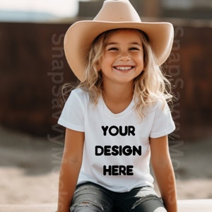 May include: A young person wearing a white t-shirt with the text "YOUR DESIGN HERE" printed on it. They are wearing a brown cowboy hat and blue jeans.