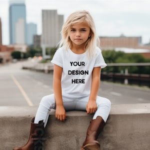 Könnte beinhalten: Ein junges Kind trägt ein weißes T-Shirt mit dem Aufdruck "YOUR DESIGN HERE" auf der Vorderseite. Es sitzt auf einer Betonkante mit gekreuzten Beinen und trägt braune Stiefel.