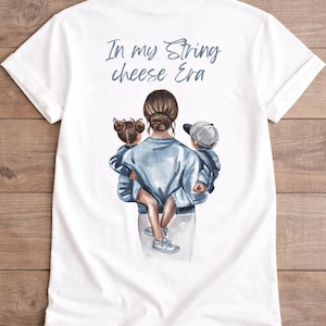 Könnte beinhalten: Weißes T-Shirt mit einer Aquarellillustration einer Person, die zwei Kinder hält. Der Text "In my String cheese Era" ist in Hellblau über der Illustration gedruckt. Das Shirt hat kurze Ärmel und einen Rundhalsausschnitt.