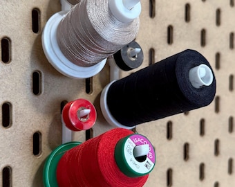 May include: Three spools of thread are displayed on a pegboard. The spools are white, black, and red. The white spool has a silver thread, the black spool has a black thread, and the red spool has a red thread. The spools are labeled with the brand name "Isacord" and the color number "1800".
