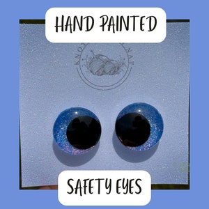 May include: Two hand-painted blue safety eyes with black pupils and glitter accents. The eyes are on a white background with the text "HAND PAINTED" and "SAFETY EYES".