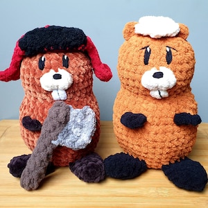 Puede incluir: Dos castores de peluche, uno marrón con un mechón de pelo blanco en la cabeza y el otro rojo y negro con un pequeño hacha en sus patas.