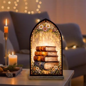 Könnte beinhalten: Dekorative Buntglaslampe mit einem Stapel von vier Büchern. Die Lampe hat einen schwarzen, bogenförmigen Rahmen mit floralen und Blatt-Akzenten. Die Bücher haben braune und blaue Einbände. Die Lampe steht auf einem Tisch mit Kerzen.
