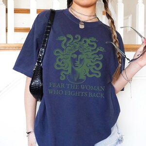 May include: A navy blue oversized t-shirt with a green graphic of Medusa, the Greek mythological figure with snakes for hair. The text "FEAR THE WOMAN WHO FIGHTS BACK" is printed below the graphic.