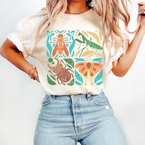 May include: A white t-shirt featuring a graphic design of four insects: a bee, a praying mantis, a beetle, and a butterfly. The insects are surrounded by green leaves and flowers. The design is in a vintage style with a distressed look.