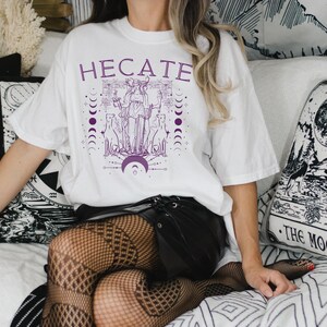 Pu&ograve; includere: Maglietta bianca con una grafica viola di una donna con tre cani e il testo "HEcate" sopra di lei.
