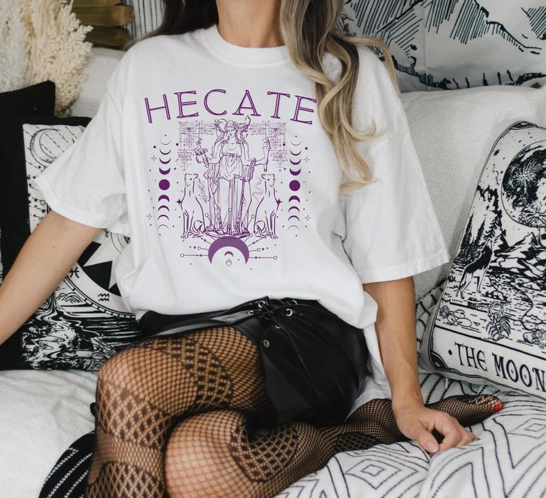 Pu&ograve; includere: Maglietta bianca con una grafica viola di una donna con tre cani e il testo "HEcate" sopra di lei.