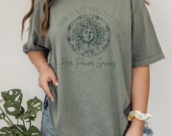 May include: A green oversized t-shirt with a graphic print of a woman's face with the text "Her Gaze Protects Her Power Grows".