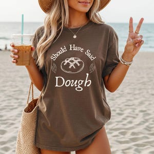 以下が含まれることがあります： 「Should Have Said Dough」の白文字とパンのイラストがプリントされた茶色のTシャツ。 ゆったりとしたフィット感で、柔らかい素材のようです。 写真はビーチで屋外撮影されています。