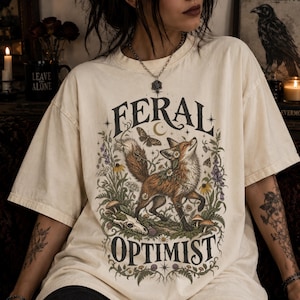 May include: An off-white short-sleeve t-shirt featuring a graphic print of a fox surrounded by wildflowers, mushrooms, a moth, and a skull. The text FERAL is printed above the fox and OPTIMIST is printed below the fox in a dark serif font.