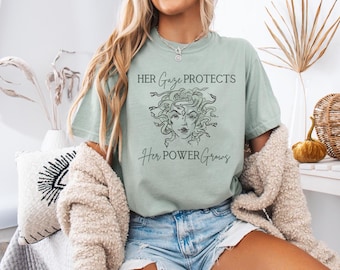 May include: A light green t-shirt with a black graphic of Medusa's head and the text "HER Gaze PROTECTS Her POWER Grows".