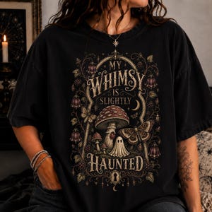 May include: A black short-sleeve t-shirt featuring a detailed graphic print with the text "MY WHIMSY IS SLIGHTLY HAUNTED". The design includes illustrations of mushrooms, a small ghost, a moth, and floral vines in muted gold and brown tones.