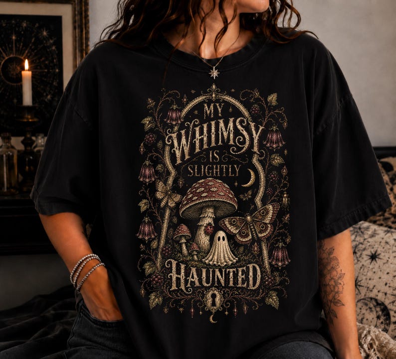 May include: A black short-sleeve t-shirt featuring a detailed graphic print with the text "MY WHIMSY IS SLIGHTLY HAUNTED". The design includes illustrations of mushrooms, a small ghost, a moth, and floral vines in muted gold and brown tones.