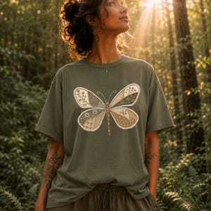 May include: Olive green t-shirt featuring a large butterfly design. The butterfly has patchwork-style wings in shades of green, beige, and brown, with floral and plaid patterns. The shirt is a casual, short-sleeved style.