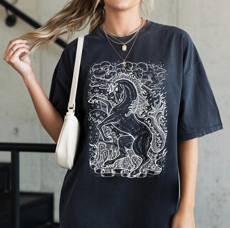 Może przedstawiać: Czarny T-shirt z białym nadrukiem jednorożca z płomieniami i gwiazdami.