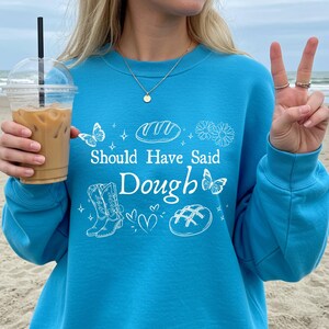 Può includere: Felpa turchese con la scritta "Should Have Said Dough" in bianco. Il design include illustrazioni di pane, farfalle, fiori e stivali da cowboy. Una tazza di plastica trasparente con caffè freddo è tenuta nella mano sinistra.