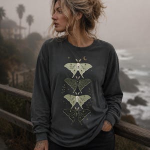 May include: A charcoal gray long-sleeve shirt featuring a vertical graphic print of four moths in varying shades of green, cream, and black. The design includes celestial elements like a crescent moon and small stars arranged between the moth illustrations.
