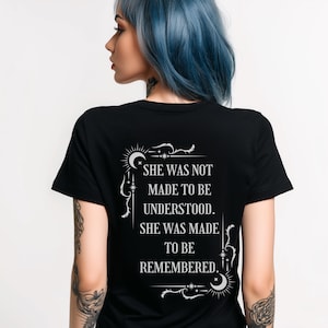 Puede incluir: Camiseta negra con texto blanco: "SHE WAS NOT MADE TO BE UNDERSTOOD. SHE WAS MADE TO BE REMEMBERED." El texto está en una fuente decorativa con acentos celestiales. La camiseta es de manga corta.