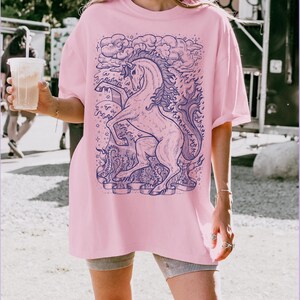 May include: A pink oversized t-shirt with a blue and white graphic of a unicorn with a cloud and stars. The unicorn is surrounded by a banner with the text "Communicate with your heart".