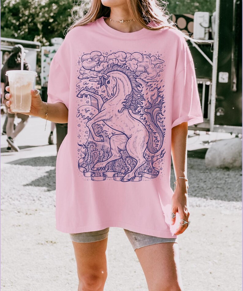 May include: A pink oversized t-shirt with a blue and white graphic of a unicorn with a cloud and stars. The unicorn is surrounded by a banner with the text "Communicate with your heart".