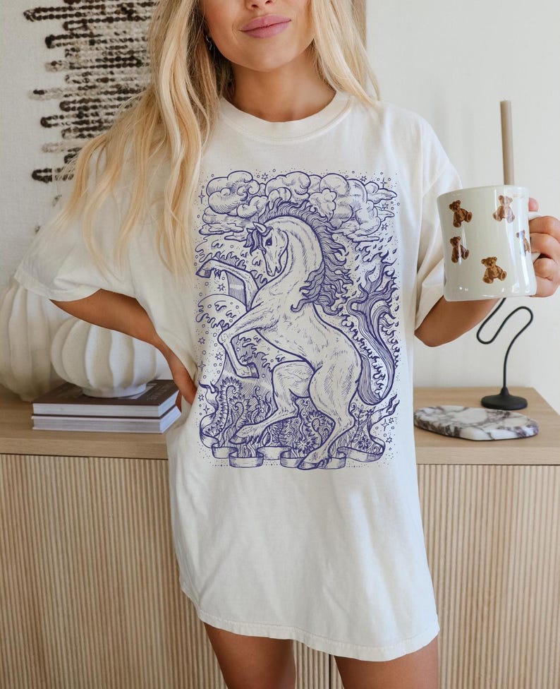 May include: A white t-shirt with a blue illustration of a unicorn with a star and clouds in the background.
