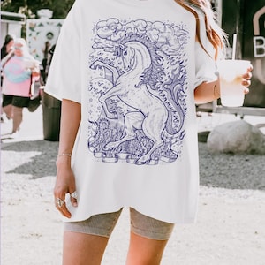 May include: White oversized t-shirt with a blue and white graphic of a unicorn with a flowing mane and tail. The unicorn is surrounded by clouds and stars.