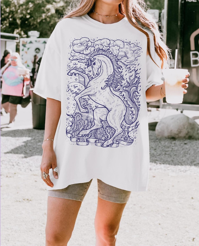 May include: White oversized t-shirt with a blue and white graphic of a unicorn with a flowing mane and tail. The unicorn is surrounded by clouds and stars.