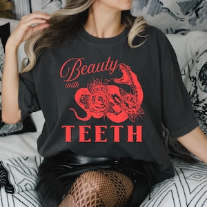 Pode incluir: T-shirt cinza escuro com um gráfico vermelho de uma cobra, rosas e as palavras "Beauty with TEETH". A camisa tem mangas curtas e um ajuste folgado. O design está centrado na frente.