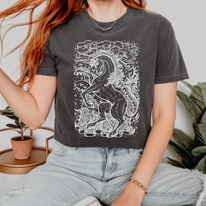 Może przedstawiać: Czarny, kr&oacute;tki T-shirt z białym nadrukiem przedstawiającym jednorożca w płomieniach. Jednorożec jest otoczony chmurami i gwiazdami.
