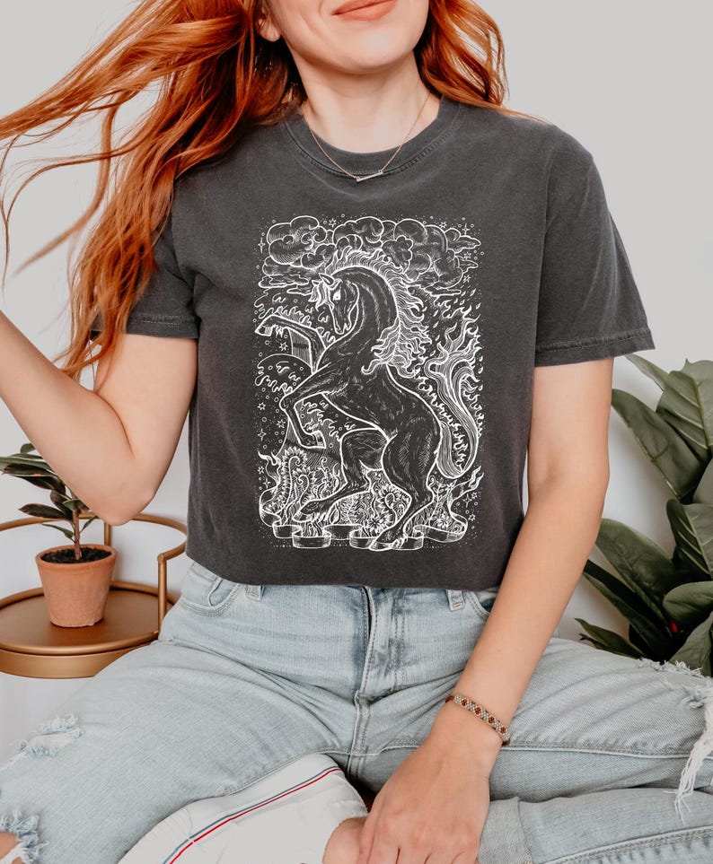 Może przedstawiać: Czarny, kr&oacute;tki T-shirt z białym nadrukiem przedstawiającym jednorożca w płomieniach. Jednorożec jest otoczony chmurami i gwiazdami.