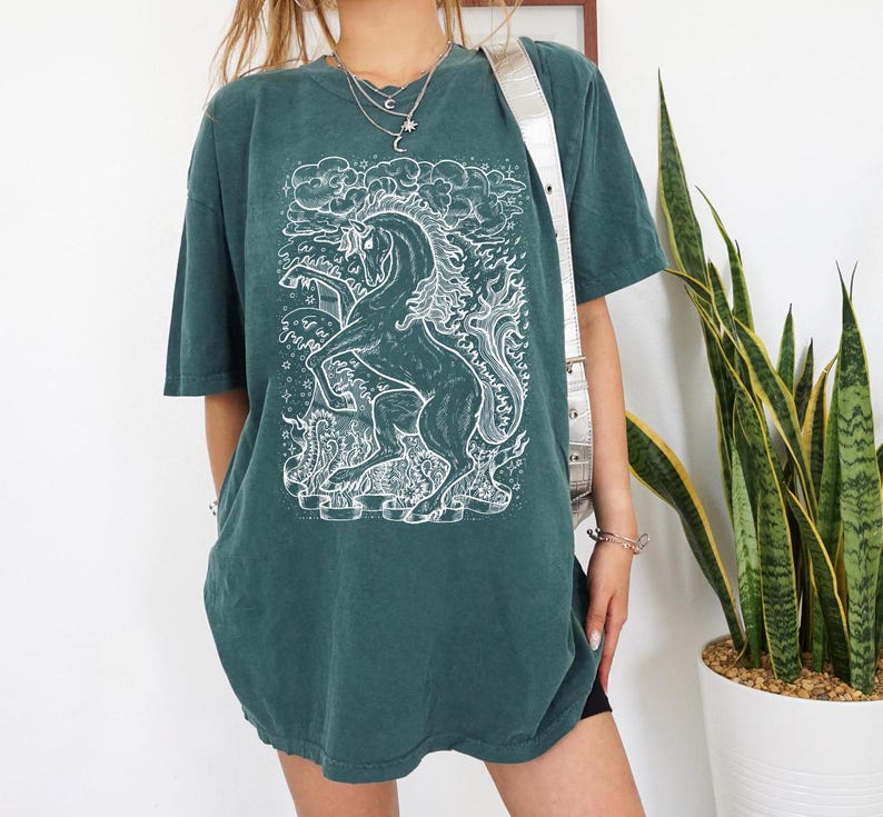 Może przedstawiać: Zielony t-shirt z białym nadrukiem konia z płomieniami i chmurami. Koszulka ma luźny kr&oacute;j i okrągły dekolt.