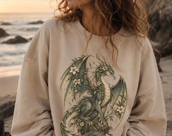 May include: A beige crewneck sweatshirt featuring a detailed graphic print of a green dragon intertwined with white flowers and foliage. The dragon is depicted in a seated position with wings partially spread, rendered in shades of green and gold.