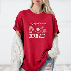 May include: A red t-shirt with the text "Loafing him was BREAD" in white. The shirt features a graphic of a scarf, bread, and leaves. The shirt is a casual style.