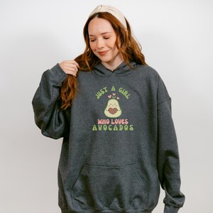 May include: Dark gray hoodie with a front pocket and a drawstring hood. The graphic on the front says "Just a Girl Who Loves Avocados" with an avocado illustration. The text and avocado are in shades of green, pink, and brown.