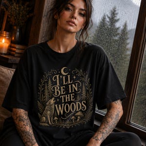 May include: A black short-sleeve t-shirt featuring a gold-toned graphic print of a howling wolf, pine trees, mushrooms, and a crescent moon. The text "I'LL BE IN THE WOODS" is centered within the design, surrounded by a decorative botanical wreath.