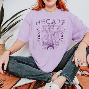 Pu&ograve; includere: Una maglietta viola con una grafica della dea greca Ecate, circondata da lune e cani. Il testo "HEcate" &egrave; stampato in viola sulla maglietta.