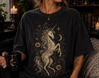 May include: A charcoal gray short-sleeve t-shirt featuring a detailed graphic print of a rearing unicorn surrounded by intricate floral patterns, stars, and a crescent moon. The design is rendered in muted gold and cream tones on the front of the shirt.