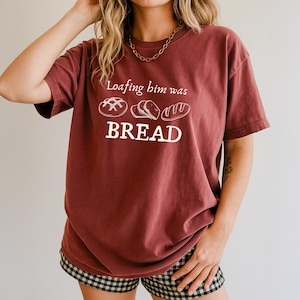 May include: A maroon t-shirt with the text "Loafing him was BREAD" in white, and bread illustrations. The short-sleeved shirt has a relaxed fit. The person is wearing black and white checkered shorts. The shirt is made of cotton.