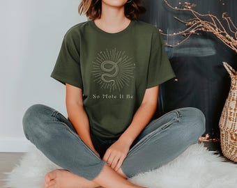 May include: Olive green t-shirt featuring a snake illustration with the text "So Mole It Be" in a light beige color. The graphic is centered on the front of the shirt. The model is wearing blue jeans.