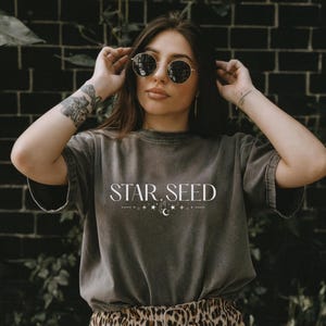 May include: A dark gray t-shirt with the text "STAR. SEED" in white, along with a celestial design. The shirt has short sleeves and a relaxed fit. The person is wearing round sunglasses.